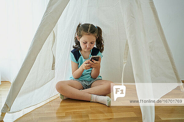 Kleines Mädchen schaut auf ihr Smartphone in einem weißen Tipi-Zelt in ihrem Haus. Technologie-Konzept