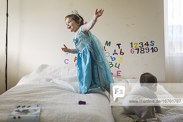 Glückliches Mädchen im Prinzessinnenkostüm  das mit seinem kleinen Bruder auf dem Bett hüpft