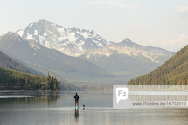 Mann auf einem Paddelbrett auf einem ruhigen See vor der Kulisse der Berge bei Sonnenaufgang in BC