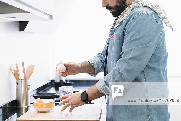 Mann im Jeanshemd beim Kochen in der Küche. Ein anonymer Mann wird ein Ei aufschlagen