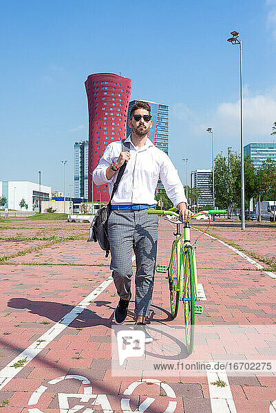 Bärtiger Mann mit Sonnenbrille  der mit seinem Fahrrad auf einem Fahrradweg im Freien spazieren geht