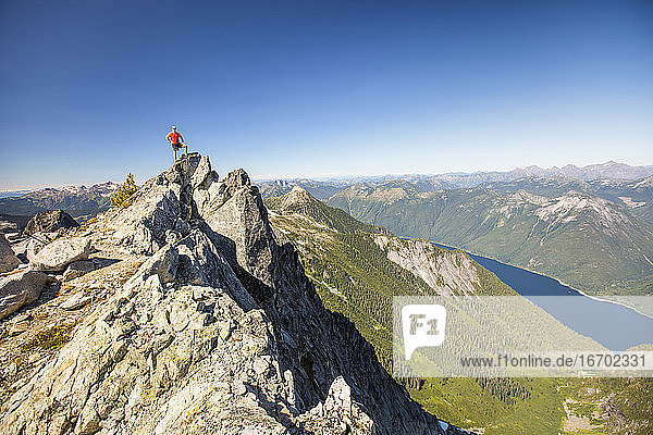 Hiker standing on mountain summit with view of lake and forest below.