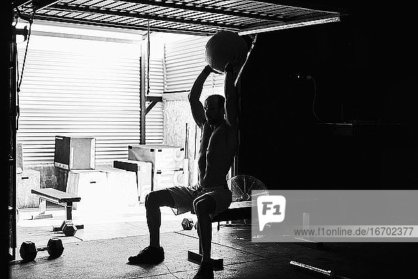 Ein fitter junger Mann trainiert mit einem Ball und Hanteln in einer Garage
