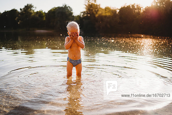 Young white boy covering face standing in water at lake golden light