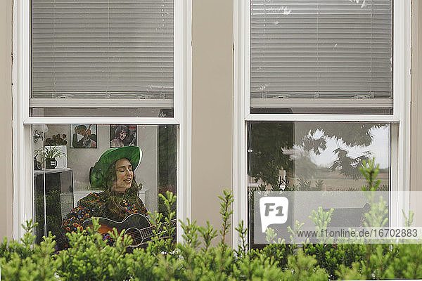 Eine Frau sitzt singend am Fenster und spielt Gitarre im heimischen Wohnzimmer