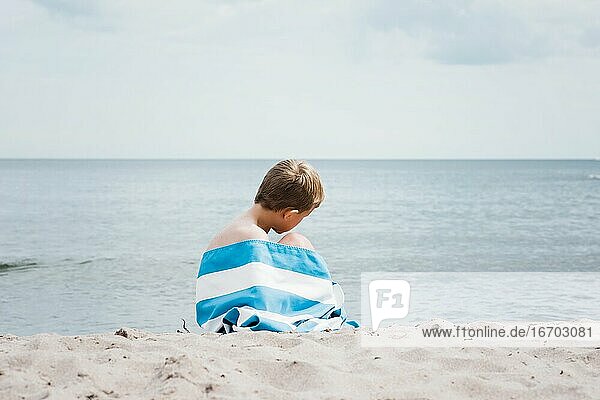 young boy sat wrapped in a striped towel alone on the beach