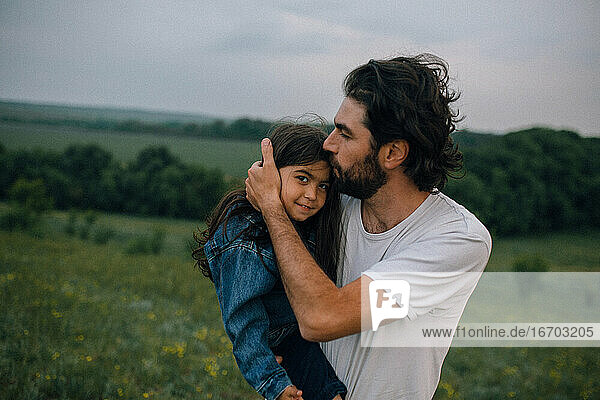 Father carrying and kissing daughter outdoors in field