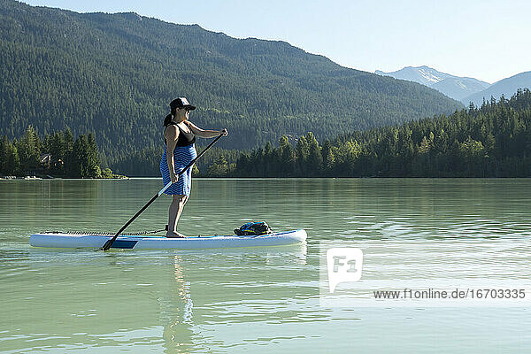 Seitenansicht einer barfuß fahrenden Frau auf einem SUP-Board auf einem See vor einem grünen Bergkamm an einem sonnigen Sommertag in British Columbia  Kanada