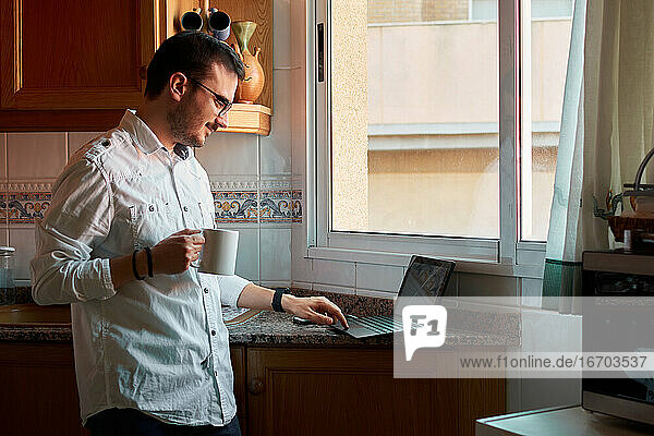 Young man looks at his laptop while drinking coffee in his kitchen