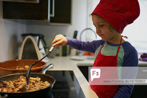 Mädchen mit roter Kochmütze brät Fleisch in einer Pfanne zu Hause
