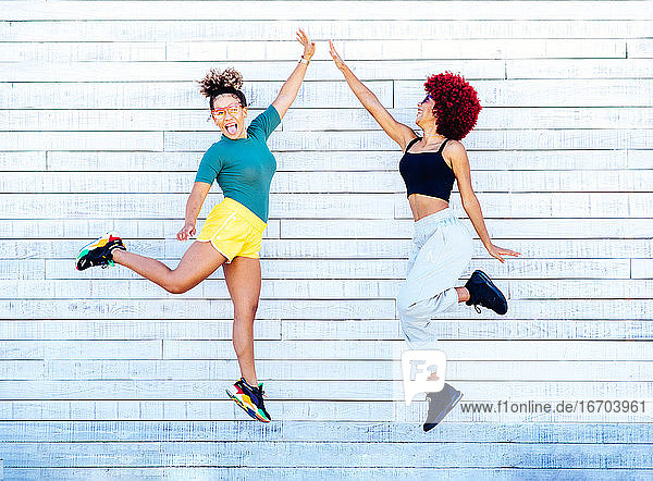 Zwei lateinamerikanische Frauen mit Afrofrisur  die auf einer weißen Wand springen und High Five machen.