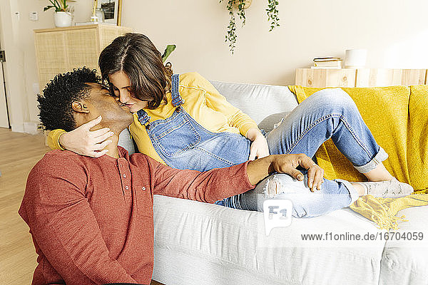 Ehemann küsst seine schwangere Frau auf einem Sofa. Interracial Paar Konzept