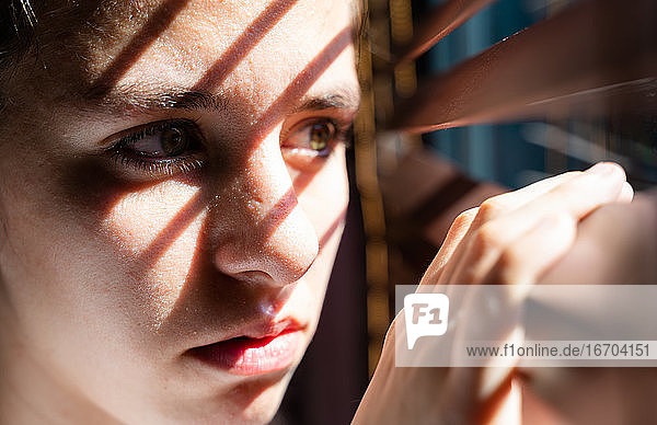 Jugendliches Mädchen mit grünen Augen öffnet einen hölzernen Vorhang mit ihren Fingern  um durch das Fenster zu schauen  während das Sonnenlicht Sonne und Schatten auf ihrem Gesicht erzeugt  das hoffnungslos erscheint. Horizontal Foto