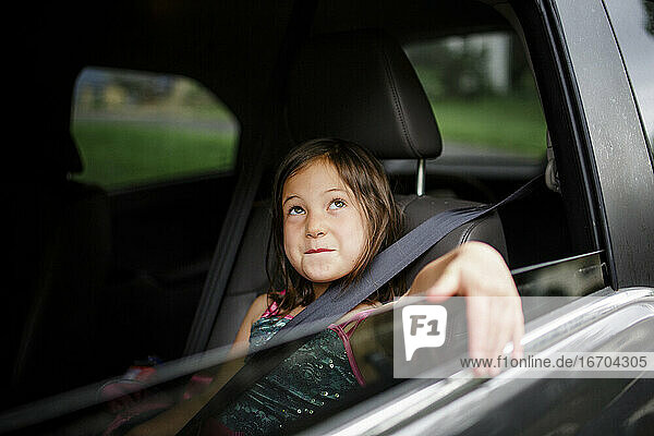A little girl sits smiling in a car with her arm draped out the window
