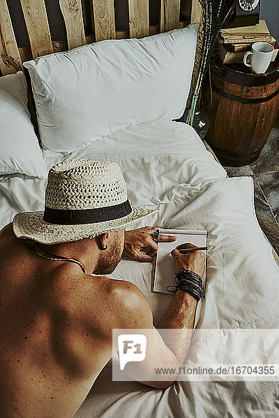 ein junger Mann mit einem Hut  der im Bett liegt und in ein Notizbuch schreibt