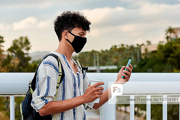 Junger afro-haariger Mann mit Maske benutzt sein Handy auf der Straße