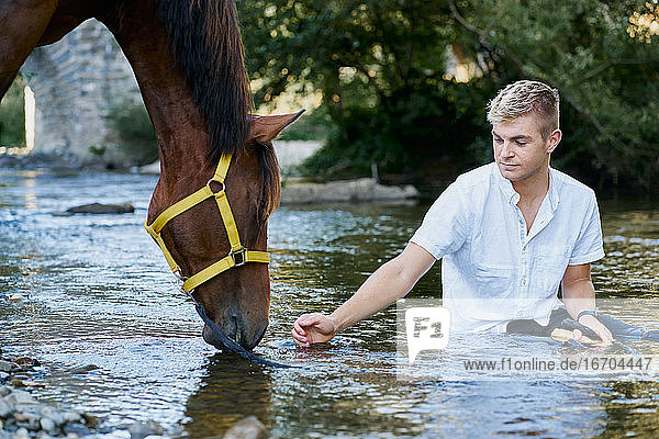 Porträt eines blonden jungen Mannes mit einem Pferd an einem Fluss