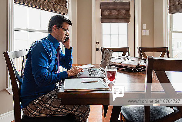 Mann am Handy  der in Pyjamahose von zu Hause aus an einem Computer arbeitet.