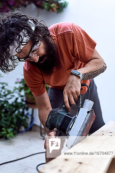 Junger Mann mit Bart  der ein Holzbrett mit einer Schleifmaschine poliert