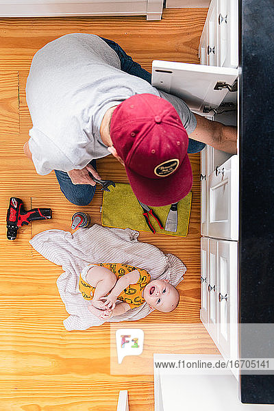 Draufsicht auf den Vater  der die Spüle repariert  während das kleine Mädchen zu Hause auf dem Parkettboden liegt