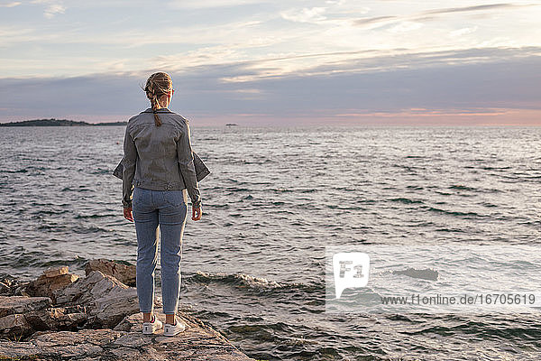 Rückenansicht einer jungen Frau  die auf einer steinernen Mole steht und auf das Meer blickt