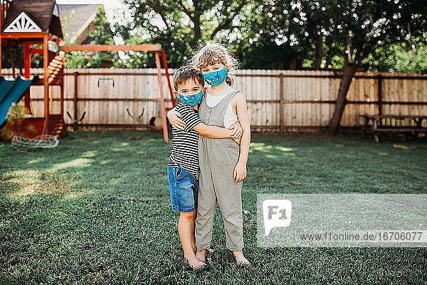 Zwei kleine Kinder umarmen sich draußen und tragen dabei selbstgemachte Stoffmasken