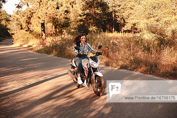Junge selbstbewusste Frau fährt Motorrad auf einer Landstraße im Wald