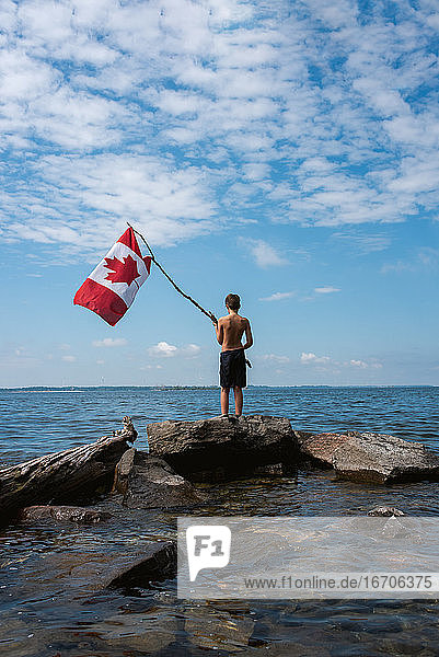 Junge hält an einem Sommertag am Ufer eines Sees die kanadische Flagge in die Luft.