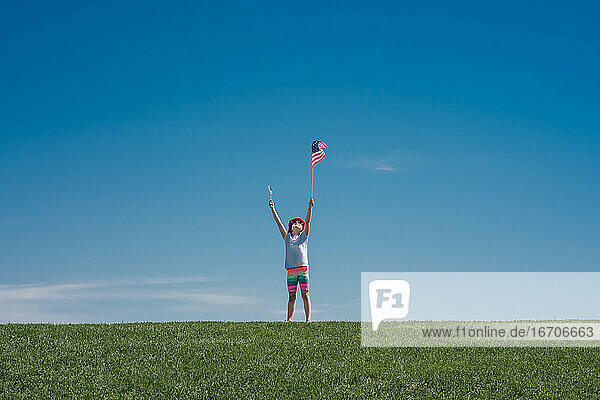 junges Mädchen hält 2 amerikanische Flaggen über ihren Kopf auf einem grünen Grashügel