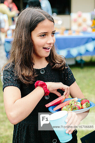 Kleines Mädchen lächelt bei einer Feier und trägt einen Teller mit Obst