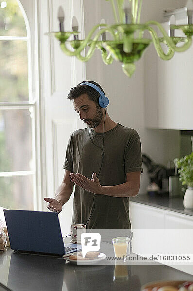 Mann mit Kopfhörern bei einem Videoanruf  der von zu Hause aus am Laptop in der Küche arbeitet