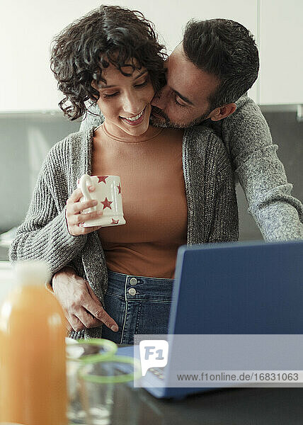 Ehemann küssen Frau arbeiten von zu Hause aus am Laptop in der Küche