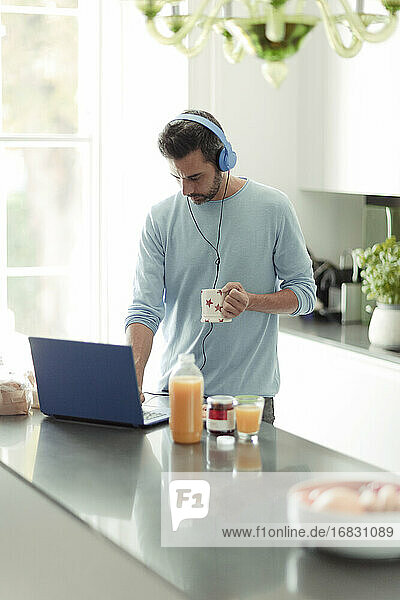 Mann mit Kopfhörern arbeitet von zu Hause aus am Laptop in der Morgenküche