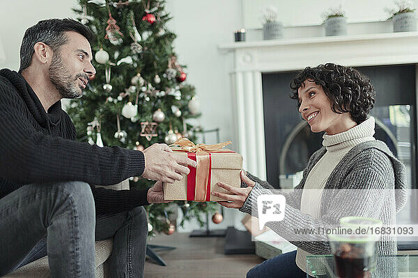 Ehemann gibt Weihnachtsgeschenk an Frau im Wohnzimmer mit Baum