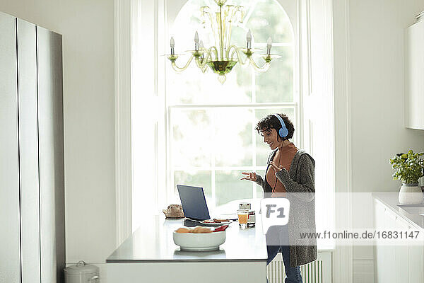 Woman with headphones video conferencing at laptop in kitchen