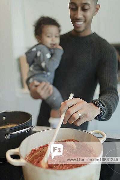 Father holding baby daughter and cooking spaghetti at stove