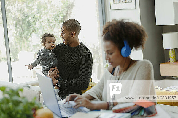 Father holding baby daughter behind working mother at laptop