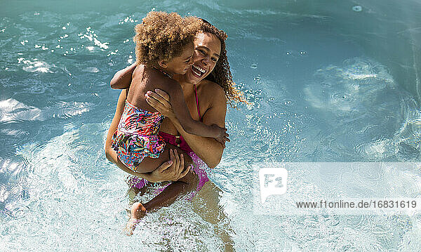 Happy mother holding daughter in sunny summer swimming pool