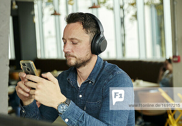 Man seated in a cafe wearing headphones  using a smart phone  working remotely.