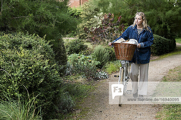 Young blond woman pushing bicycle with basket along footpath.