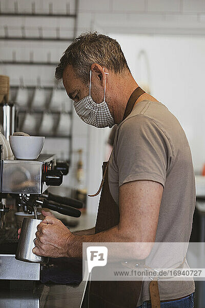 Männlicher Barista mit brauner Schürze und Gesichtsmaske arbeitet in einem Café und schäumt Milch auf.