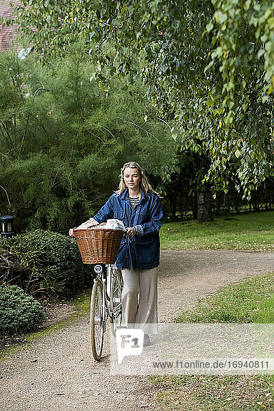 Junge blonde Frau  die ein Fahrrad mit Korb auf dem Fußweg schiebt.