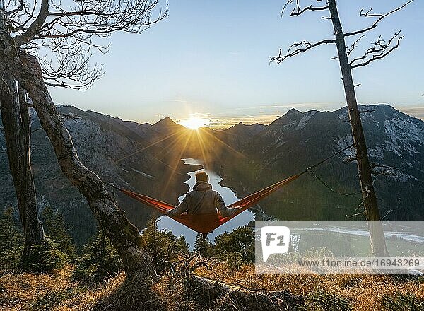 Junger Mann sitzt in einer roten Hängematte  Panoramablick auf Berge mit See  Sonnenstern  Sonnenuntergang  Plansee  Tirol  Österreich  Europa