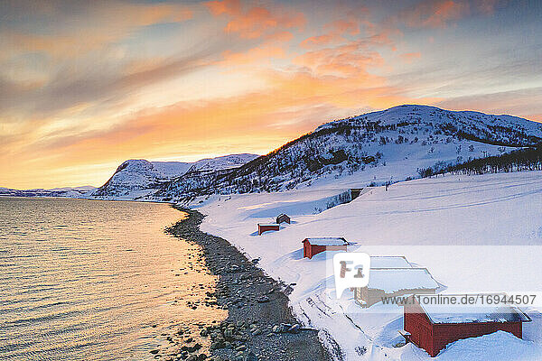 Sonnenuntergang über roten Hütten im Schnee am Porsangerfjord mit Nordkap (Nordkapp) im Hintergrund  Troms og Finnmark  Arktis  Norwegen  Skandinavien  Europa