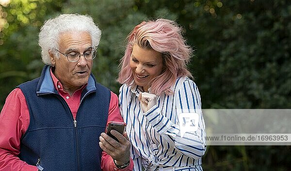 Eine hübsche 34-jährige blonde Frau  die sich mit einem 67-jährigen Mann unterhält und dabei auf ein Mobiltelefon schaut.