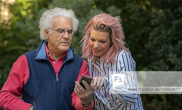 Eine hübsche 34-jährige blonde Frau  die sich mit einem 67-jährigen Mann unterhält und dabei auf ein Mobiltelefon schaut.