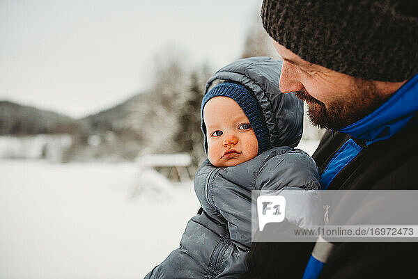 close up of Dad holding baby wearing snowsuit in winter with snow