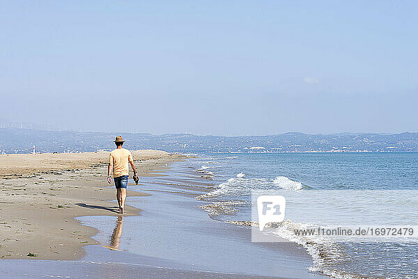 Rear view of man with hat  holding shoes walking barefoot on seashore