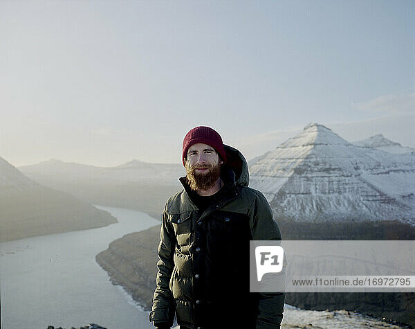 Man looking at camera on snowy mountain toop looking towards the ocean in the Faroe Islands