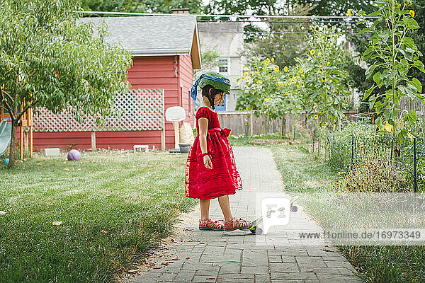 Ein Mädchen in rotem Kleid und Helm Schritte auf Skateboard im Garten im Sommer